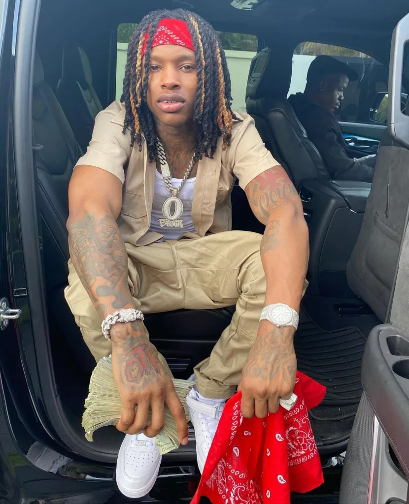 King Von sitting in his car and holding cash and red bandana in hand.