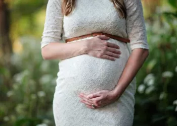 A pregnant lady posing with her baby bump wearing a white dress.
