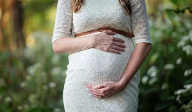 A pregnant lady posing with her baby bump wearing a white dress.
