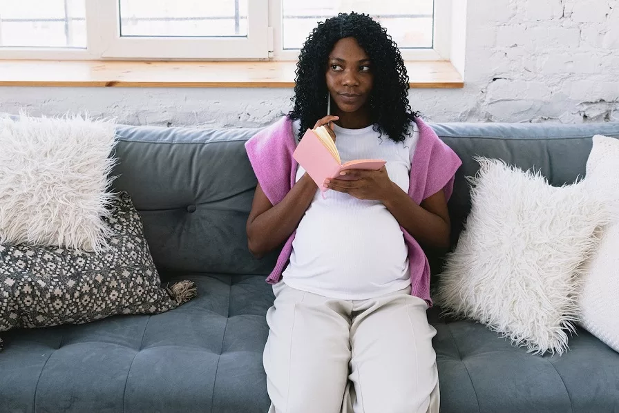 Pensive black pregnant woman writing in notebook on sofa