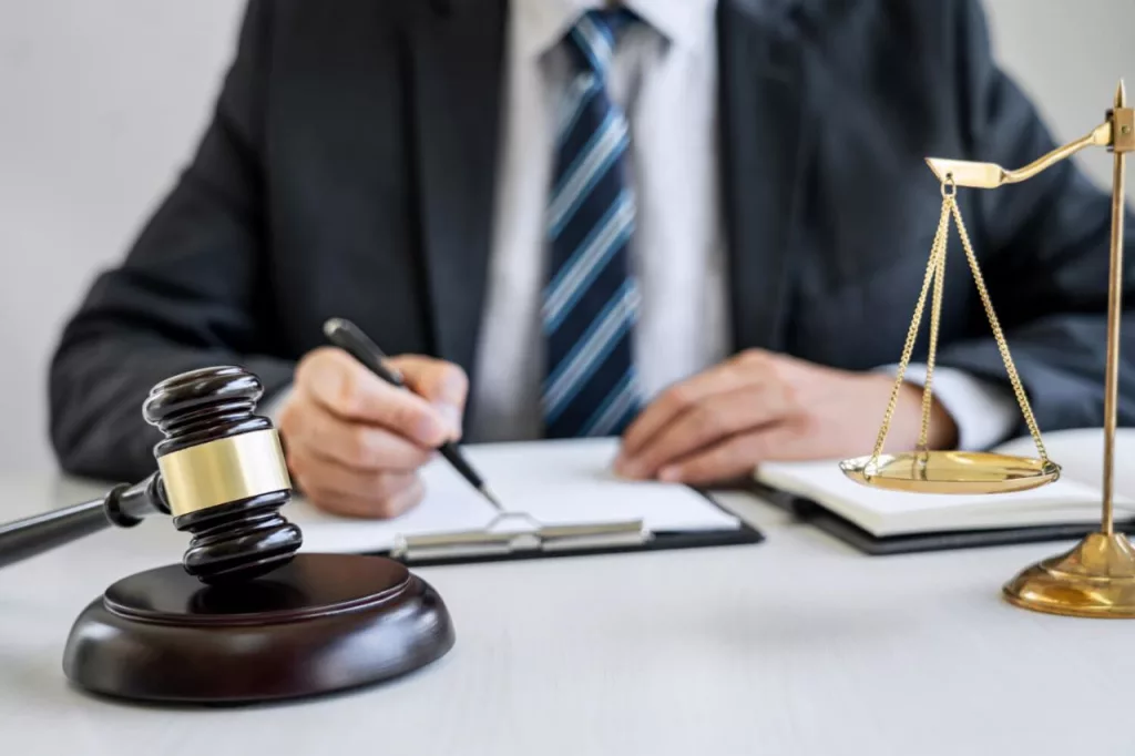 A person writing in a diary. A gavel and justice scales are also placed on the same table.