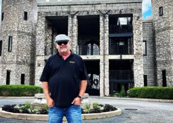Rick Harrison wearing black polo t shirt, blue jeans and grey cap. He is posing outside the Kentucky Castle.