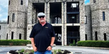 Rick Harrison wearing black polo t shirt, blue jeans and grey cap. He is posing outside the Kentucky Castle.