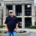 Rick Harrison wearing black polo t shirt, blue jeans and grey cap. He is posing outside the Kentucky Castle.