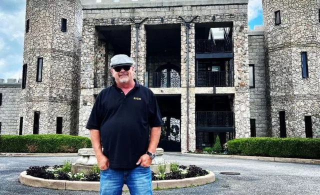 Rick Harrison wearing black polo t shirt, blue jeans and grey cap. He is posing outside the Kentucky Castle.