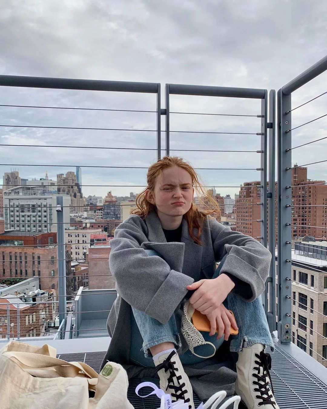 Sadie sitting on the roof top wearing grey colored hood and blue jeans.