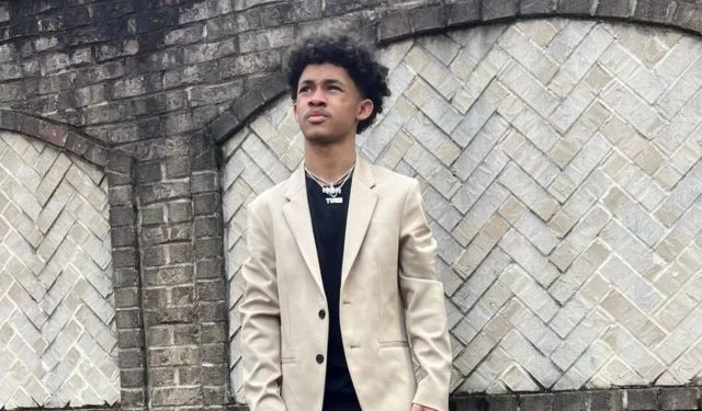 Dwayne wearing black shirt and jeans with light colored coat and sports shoes.