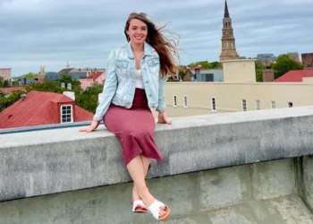 Taelyn sitting on the terrace wall of a building. She is wearing red skirt, white top, sky blue jacket and white footwear.