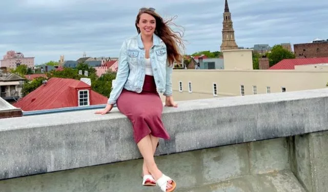Taelyn sitting on the terrace wall of a building. She is wearing red skirt, white top, sky blue jacket and white footwear.